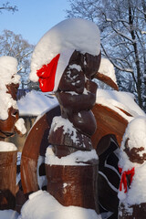 Wooden sculpture covered with fresh snow in winter park. Carved wooden figure wearing a red winter hat, surrounded by snow-covered trees under clear blue sky. Seasonal outdoor scene, winter weather