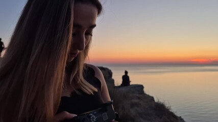 Woman camera sunset golden hour ocean view. Female photographer adjusting settings with scenic seascape and warm sky background.