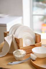 Set of white dishes with cleaning powder and brush on table in kitchen, closeup