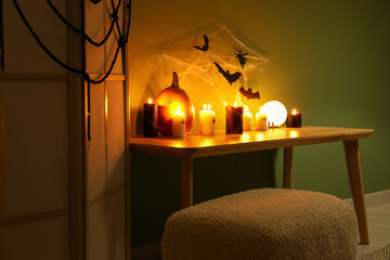 Table with burning candles, folding screen, bench and Halloween decor near green wall in dark room