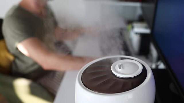 Modern air humidifier device during sitting workplace, young man using computer work on internet, enjoying aromatherapy steam scent from essential oil diffuser in room at home office.