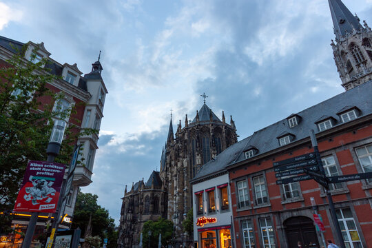 Evening street view of the Gothic choir of Aachen Cathedral in Germany. The scene includes historic city buildings, a directional signpost for tourists, and the illuminated windows of local shops