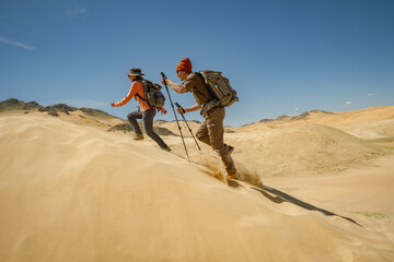  Two hikers with backpacks and trekking poles running or climbing up a steep sand dune under a bright blue sky in an arid desert landscape. © ANDREY