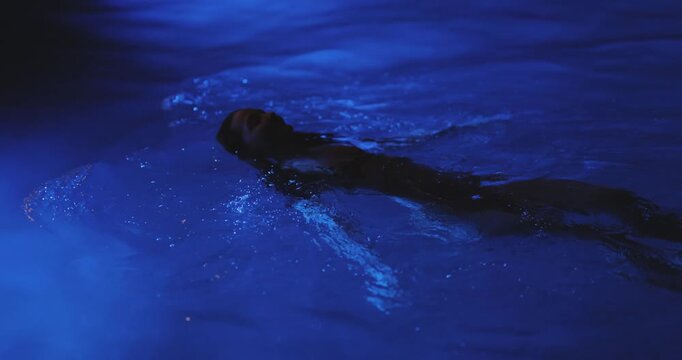 Woman Swimming Alone In Pool At Night With Moody Blue Lighting