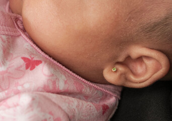 Fototapeta premium Extreme close-up shot focusing on the delicate ear of an infant wearing a small, golden stud earring with a green stone. The baby's skin texture is visible, resting against soft pink fabric patterned