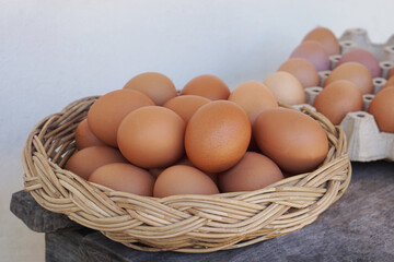 Fresh brown chicken eggs arranged in a wicker basket on a rustic wooden surface, with egg trays in the background. Natural organic food concept, farm fresh ingredients, healthy protein and agriculture