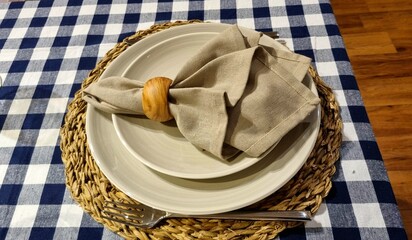 Elegant  home table setting, classic tablecloth with a blue and white check, wicker round stand and  beige plates. A napkin decorates a plate nature texture with a wooden ring, and cupronickel utensil