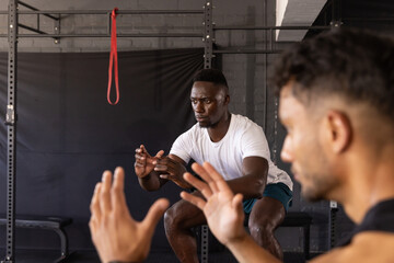 Diverse male workout partners squatting in gym with squat rack, red resistance band, weight bench