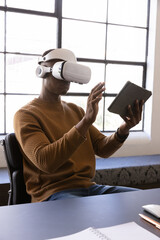 Man wearing white over-ear vr headset interacting with dark tablet at desk near grid window © wavebreak3