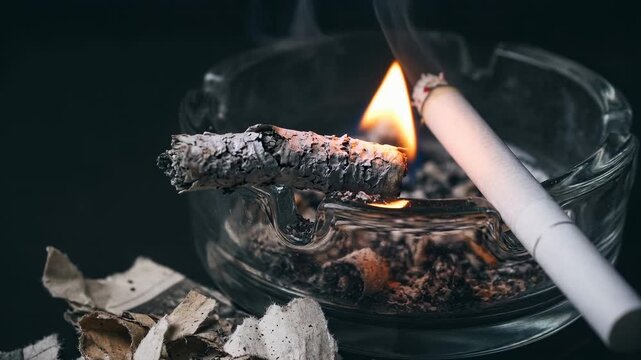 A close-up shows a glass ashtray filled with wet ashes and burnt paper. A cigarette smolders, releasing smoke and heat in a dimly lit room.