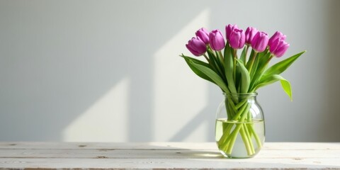 A bouquet of vibrant purple tulips arranged in a clear glass vase sits on a rustic white wood surface, bathed in soft, natural light