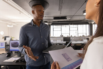 Diverse coworkers discussing printed bar-chart reports at desk in open plan office with laptop