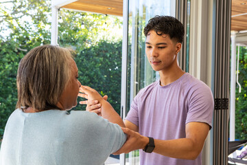 Senior Hispanic woman stretching arms as Asian man guiding movement on home patio with glass doors