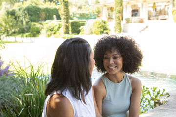 Diverse mother and daughter chatting and sitting on stone ledge by water feature in garden