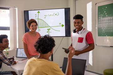 Diverse teacher and students analyzing cosine graph on monitor at table in classroom with laptop