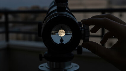 Teen observing the moon through a telescope on a balcony at dusk, with a hand adjusting the focus, cityscape in the background, looking