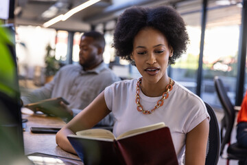 African american coworkers reading hardcover book and using tablet at office desks next to windows