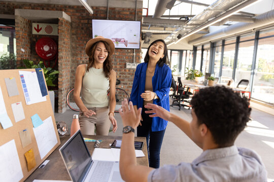 Diverse coworkers laughing, celebrating milestone at design studio desk with laptop and pinboard - Powered by Adobe