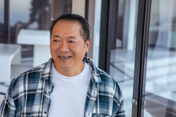 Asian senior man smiling while glancing left on terrace beside bench and sliding glass doors