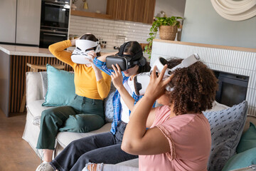Naklejka premium Diverse female friends wearing VR headsets while sitting on sofa among patterned cushions at home