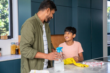 Indian father with son standing at countertop in home kitchen spraying cleaner while wearing gloves © wavebreak3