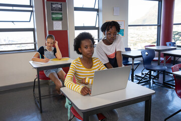 Diverse female classmates collaborating on laptop and writing in notebook in classroom