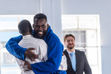 Diverse male judo partners embracing at dojo with blue, white gis, belts, suit, tie