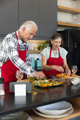 Senior couple in red aprons slicing vegetables on kitchen island with chopping boards and olive oil