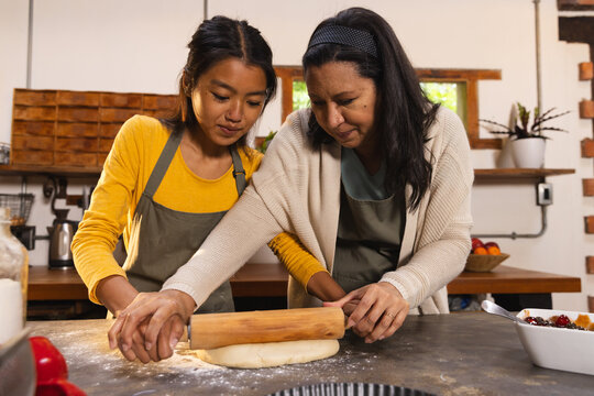Diverse mother and teenage daughter wearing aprons rolling dough with wooden pin in home kitchen