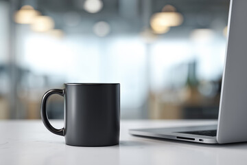 Fototapeta premium A black ceramic mug stands on a white table next to a silver laptop against a blurry background of a modern office