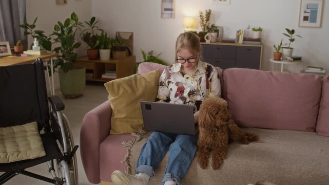 Smiling gen Z girl with paraparesis working from home on couch with cute miniature poodle lying near her