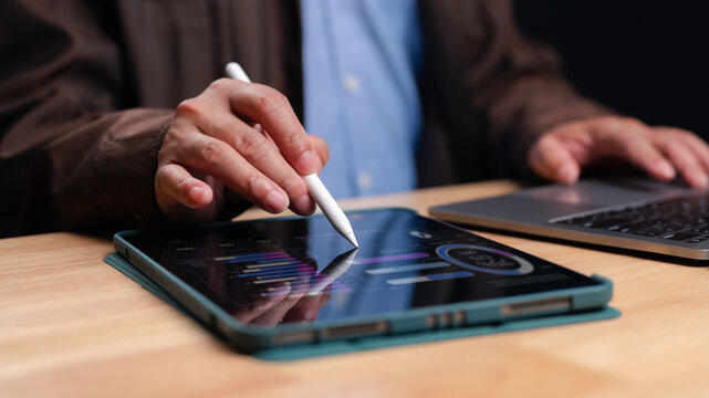 Professional businessman analyzes financial data reports using digital tablet stylus. Modern office workspace features laptop computer wooden desk surface. Detailed charts graphs display market