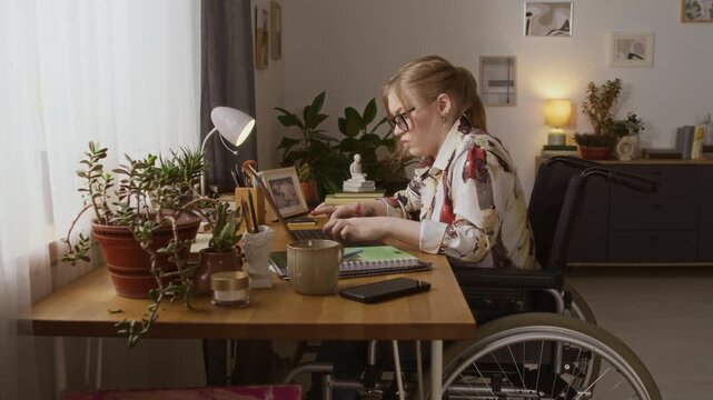 Zoom in side view of Caucasian girl with paraplegia typing on laptop while sitting in wheelchair at wooden desk at home