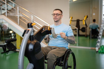 Focused disabled man in wheelchair using hand cycling machine in gym at training session, spinning...
