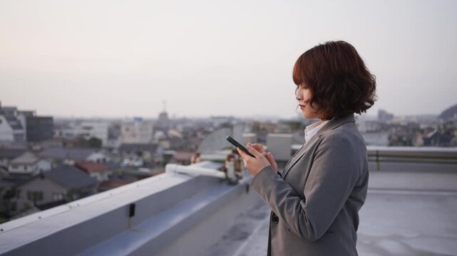 夕暮れのビル屋上でスマートフォンを操作し物思いにふける若い会社員女性