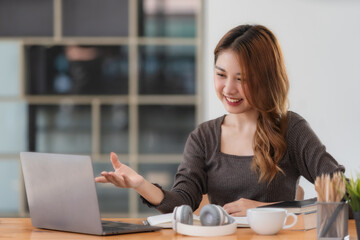 An attractive Asian woman is making a video call with a laptop computer and waving her hand. Young females communicate with family and friends online at home, online E-learning and studying.