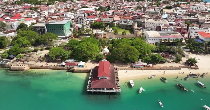 Cinematic aerial drone flight over Forodhani Gardens and the Old Fort in Stone Town, Zanzibar. Oceanfront park, historic Swahili architecture, and vibrant coastal life create a stunning travel scene.