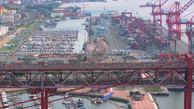Side-on cinematic aerial drone shot of half marathon and 10K long-distance running event in Lisbon, Portugal, Europe. Runners crossing the red 25th April bridge