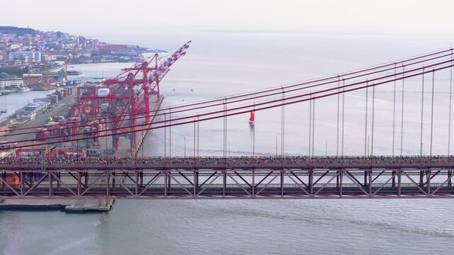 Side-on aerial drone shot of half marathon and 10k runners crossing the iconic red 25th April suspension bridge above the Lisbon waterfront, marina, port docks, and industrial cranes in Portugal
