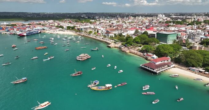Cinematic forward-moving drone shot flying over boats toward Forodhani Gardens in Stone Town, Zanzibar on a sunny summer day. Turquoise water, coastal life, and historic skyline create a vibrant trave