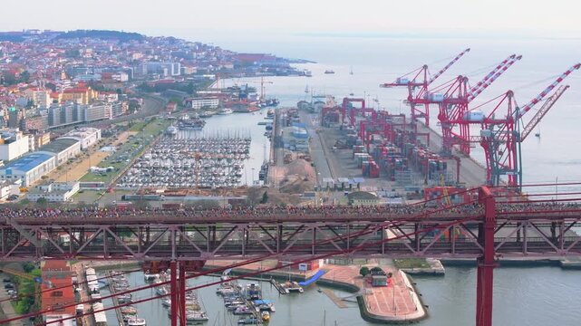 Aerial side-on drone view of thousands of half marathon and 10k runners crossing the red 25th April suspension bridge beside the Lisbon waterfront, marina, port docks, industrial cranes in Portugal