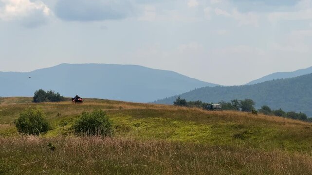 Off-road vehicle parked on a wide mountain meadow with layered forested hills and distant mountains in the background. Scenic rural landscape with open space, calm atmosphere, and natural light.