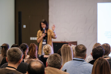 Obraz premium Bright speaker presents at conference. Audience listens attentively. Woman holds microphone, raises hand. Modern meeting space with projector screen. 
