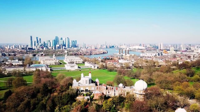 Aerial drone shot elevating above Royal Greenwich Observatory, revealing Isle of Dogs and Canary Wharf skyline, modern skyscrapers, sunny day, blue sky, cinematic London cityscape