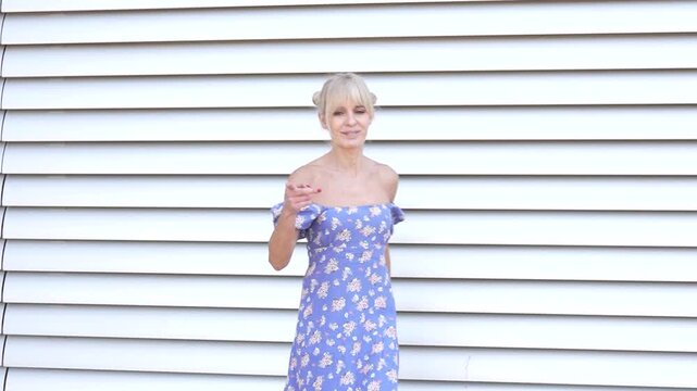 Blonde woman in blue summer dress posing outdoors against white wall, emanating carefree confidence