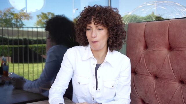 A stylish woman sits in a cozy cafe, showing off her curly hair with a thoughtful look