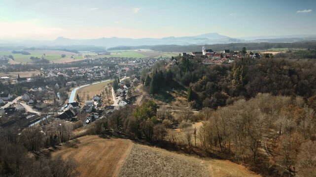 Sweeping backward drone pullback from Aach village along river valley, highlighting hillside homes, church, autumn woods, fields, and expansive mountain backdrop. Perfect for travel docs, regional ads