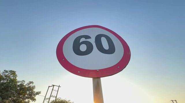 Low-angle orbiting shot around circular 60 speed limit sign, bold numerals clear against blue sky, sunlight flaring softly as camera circles roadside pole on open highway