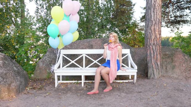 Blonde girl sit on bench and lick sweet candy, pack balloons float in air, tied to seat arm. Positive young woman rest alone at park, enjoy lollipop. Soft evening lighting, tender colours