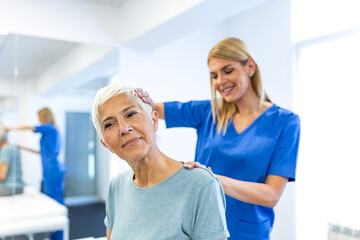 Obraz premium Professional female physiotherapist in blue scrubs performing neck mobilization and cervical spine stretching on a senior woman to improve range of motion in a bright rehabilitation clinic center.